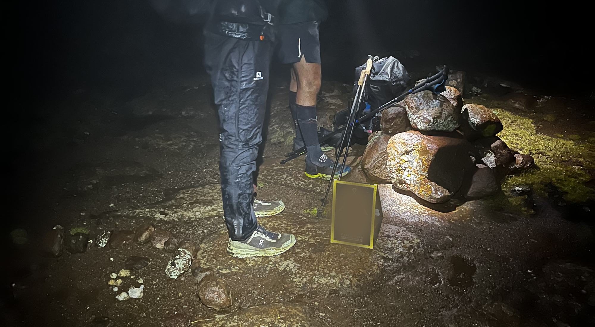 Two runners stopped at a summit cairn in the dark, headlamps on wet rock